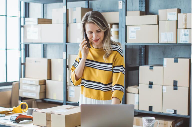 Smiling woman standing in warehouse checking laptop while on the phone