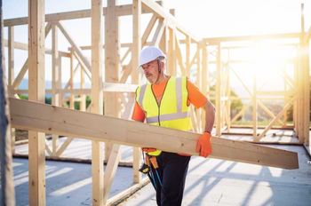 Man wearing construction helmet and high visibility vest carrying wood beam in construction site