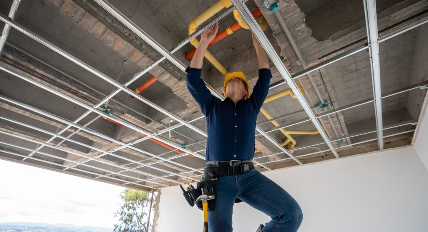 Man on ladder wearing construction helmet repairing pipes in commercial building ceiling