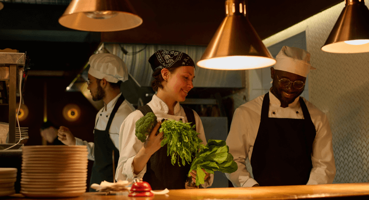 Chef in low lit kitchen holding fresh greens standing next to coworker prepping food