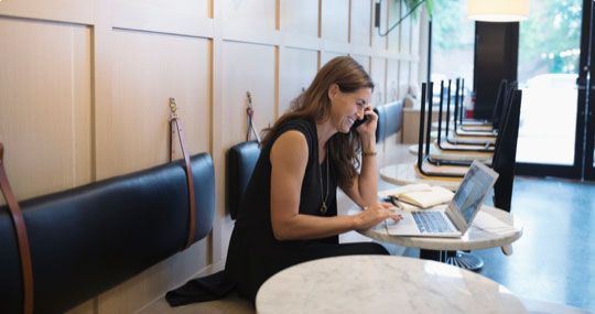 Smiling woman with laptop open sitting at table in cafe while taking phone call