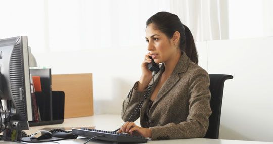 Professionally dressed woman sitting at desk typing on keyboard while taking phone call