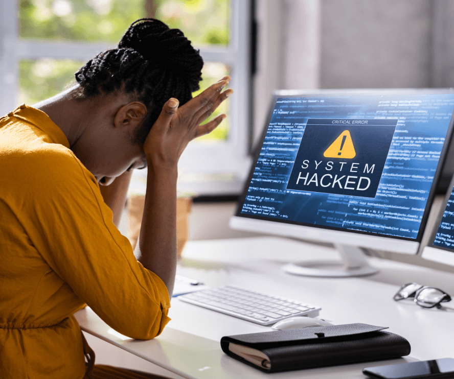 Distressed woman sitting at desk with system hacked message on computer monitor in front of her