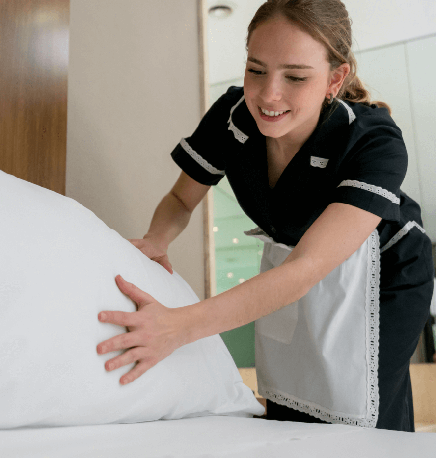 Woman in hotel maid uniform fluffing pillow on freshly made bed