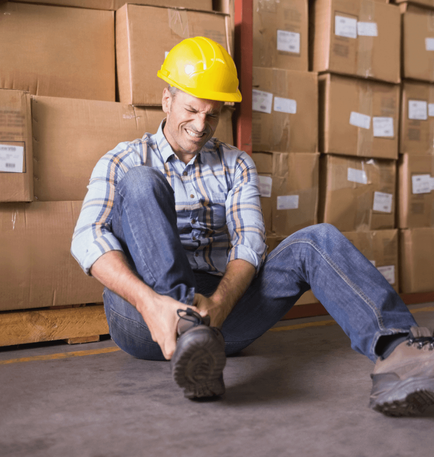 Man in hardhat sitting on ground in warehouse grasping injured ankle