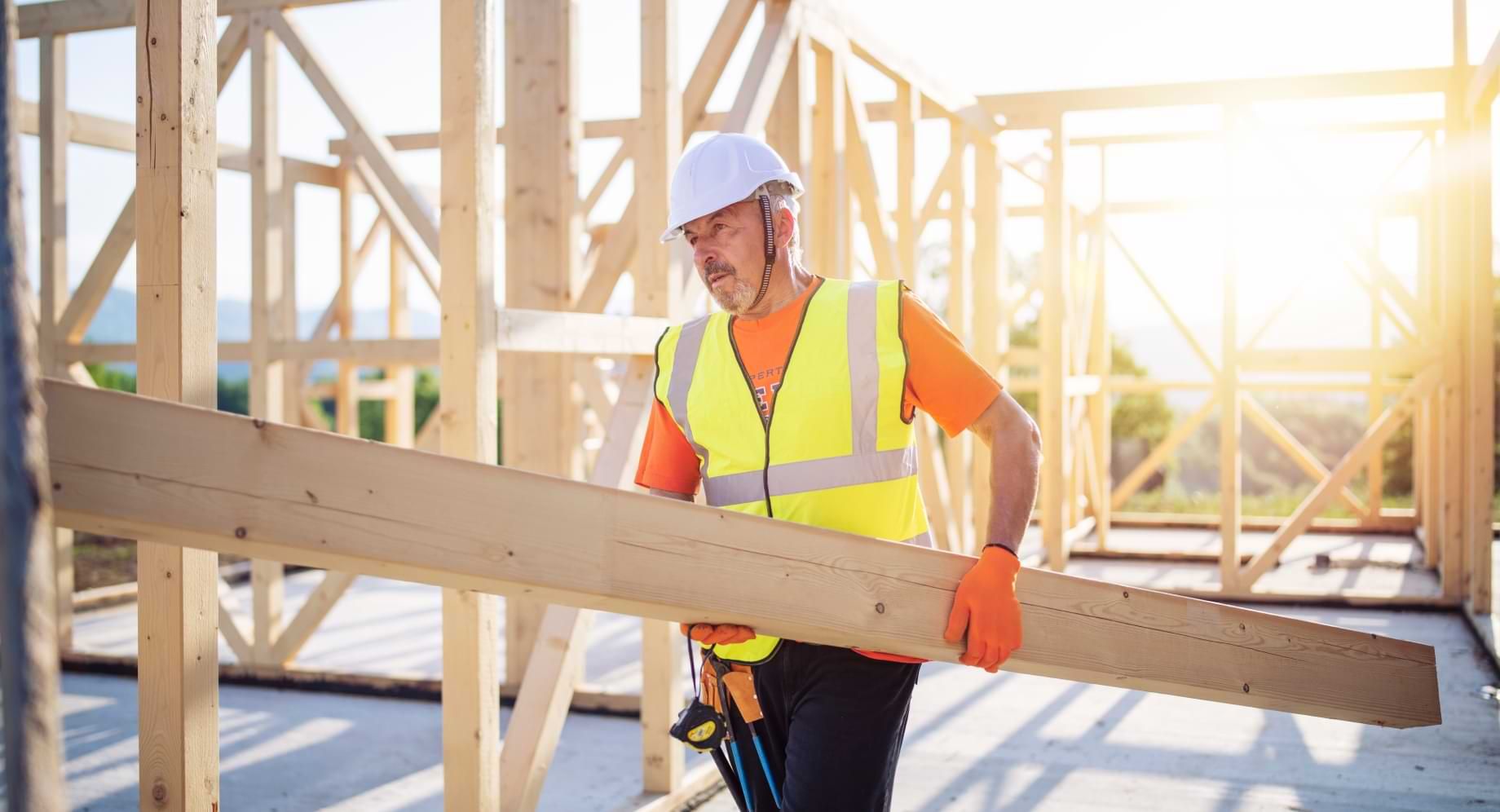 Man wearing construction helmet and high visibility vest carrying wood beam in construction site